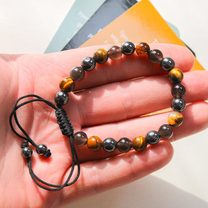 Close-up of the Azenway Triple Protection Bracelet held in the hand, showing polished Tiger’s Eye, Black Obsidian and Hematite beads with a woven adjustable cord.