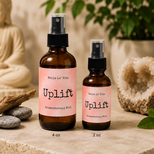 Bella La’ Vita Uplift Aromatherapy Mist in 4 oz and 2 oz amber glass spray bottles displayed on a warm, spa-inspired backdrop. The soft peach labels feature the scent name "Uplift" in a minimalist design. The bottles are styled with smooth river stones, natural driftwood, a sparkling crystal geode, fresh green leaves, and a softly blurred Buddha statue, creating a bright, uplifting atmosphere that inspires positivity and everyday wellness.