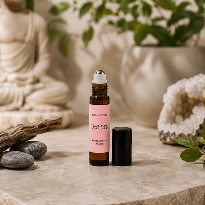 Bella La’ Vita Uplift Aromatherapy Roller in an amber glass roll-on bottle with the cap removed to reveal a stainless steel rollerball applicator. The soft peach label features the scent name "Uplift" in a minimalist design. Displayed on a warm, spa-inspired backdrop with smooth river stones, natural driftwood, a sparkling crystal geode, fresh green leaves, and a softly blurred Buddha statue, creating an uplifting atmosphere that evokes positivity, energy, and everyday wellness.