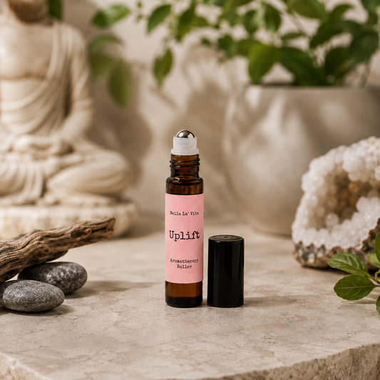 Bella La’ Vita Uplift Aromatherapy Roller in an amber glass roll-on bottle with the cap removed to reveal a stainless steel rollerball applicator. The soft peach label features the scent name "Uplift" in a minimalist design. Displayed on a warm, spa-inspired backdrop with smooth river stones, natural driftwood, a sparkling crystal geode, fresh green leaves, and a softly blurred Buddha statue, creating an uplifting atmosphere that evokes positivity, energy, and everyday wellness.
