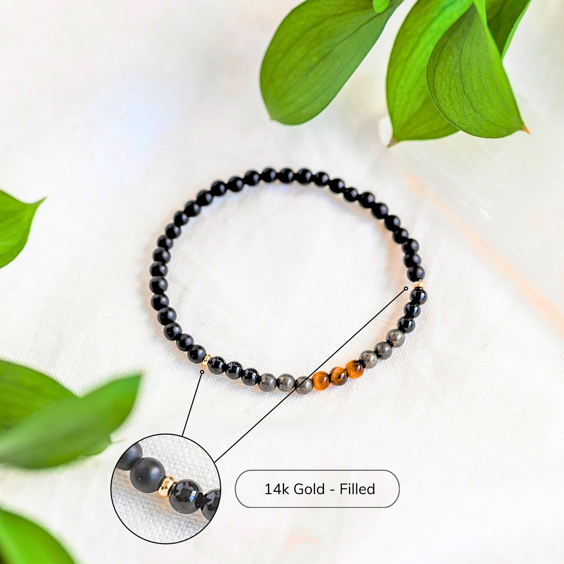 Protection bracelet with Black Tourmaline, Black Onyx, Pyrite and Tiger’s Eye featuring 14k gold-filled beads shown on white cloth with greenery