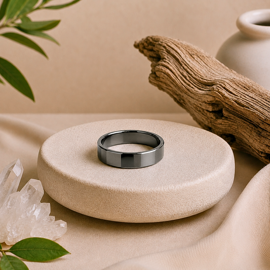 Polished hematite crystal ring with a sleek metallic gray finish and faceted band displayed on a textured cream stone pedestal, surrounded by clear quartz, driftwood, greenery, and soft beige fabric.